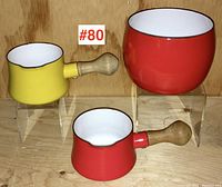 Three-piece Dansk enamelware set including large red bowl and two small handled pots, one red and one yellow, displayed on clear acrylic risers against wood background.