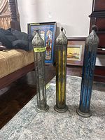 Three tall vertical lanterns in clear, amber, and blue glass with pierced tin tops and bases placed on floor carpet.