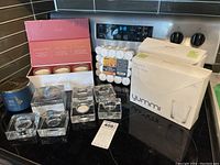 Lot items arranged on black stovetop countertop: glass votive holders, Illume scented candles box, 32-pack tea lights, blue jar candle.