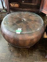 Round leather coffee table with brown textured leather, thick round beveled glass top and dark carved wooden footed base, viewed from slightly above.