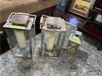 Photo showing two large silver-tone metal framed glass lanterns with wood handles, one including decorative stones, a white pillar candle, and battery lights, next to a smaller wooden framed lantern with a tin roof and green star design on glass