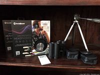 Two boxed SoundBound wireless stereo headphones, two pairs of Bushmaster mini binoculars with cases, small tripod with telescoping legs placed on a dark wood shelf.
