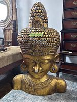 Front view of large gold-colored Buddha bust sculpture with intricate headdress and facial features, showing size in relation to nearby furniture.