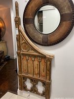 Side view of the decorative wooden panel showing carved Gothic arches and painted detailing leaning against a wall beneath a round leather and wood mirror.