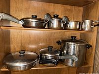 Photo showing multiple pieces of RevereWare cookware including pots and pans with stainless steel bodies, black handles and lids, placed on shelves.