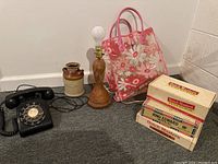Photo showing the entire lot: black rotary dial telephone, wooden lamp with bulb, ceramic milk can labeled 'MILK', pink floral plastic tote bag, and three vintage cigar boxes.