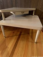 Full front view of the MCM two-tier corner table showing triangular stepped surfaces and drawer with brass handle, tapered legs with brass caps.