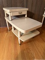 Front angled view showing the two-tier stepped design, drawer panel with brass handle, tapered legs and top shelf surface.