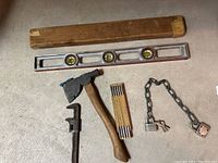 Group of older tools displayed on floor: wooden level, hatchet, folding ruler, caliper, metal chain, and two padlocks with keys.