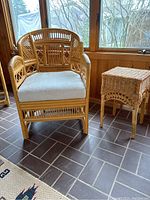 Front view of rattan chair with light fabric cushion and rattan side table on tile floor next to windows.