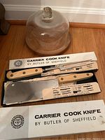 Photo of cutlery set with Carrier Cook Knife and Fork by Butler of Sheffield in original boxes, and glass dome cheese tray with wooden base behind them.