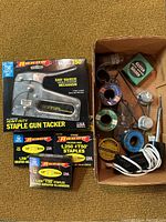 Photo showing soldering tools and materials in a cardboard box, including soldering iron with cord, spools of solder wire, flux bottle, and other small soldering accessories.