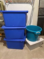 Photo showing three stacked large blue plastic storage totes, one clear storage bin with white lid, and one round teal bin with white rope handles.