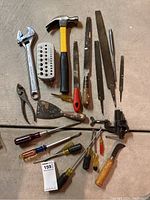 Group of hand tools including wrench, hammer, files, screwdrivers, putty knife, pliers, and metal grater-like tool spread on floor