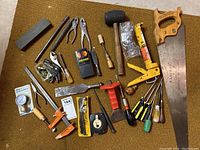 Photo showing multiple hand tools and hardware on carpet including sharpening stone, rubber mallet, hand saw, screwdrivers, clamps, and small hardware parts.