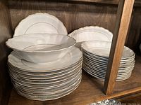 Stack of twelve dinner plates with a ribbed texture and gold trim, several oval platters stacked behind.