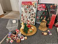 Wooden Christmas ornaments in packaging, lighted musical bells, several figurines, tiny wrapped presents, and gold charger plate arranged together on floor.