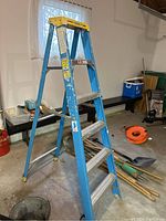 Blue Werner fiberglass A-frame ladder standing open in a garage with visible wear, scuffs, and paint marks.