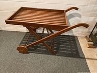Wooden cart with slatted top, foldable X-shaped base, two small round wooden wheels at front, curved wooden handles, medium brown wood finish, placed on grey carpet in front of a white wall.