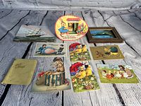 Multiple Easter-themed vintage postcards, two small paintings on board, and small pale yellow photo album displayed on wooden surface.