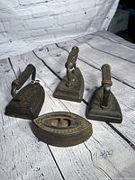 Four antique cast irons arranged on a wooden surface against a brick wall background, showing different shapes and wooden handles.