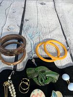 Photo showing two wooden bracelets, two yellow plastic bangles, green carved brooch, and some earrings and necklaces on black display board.