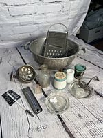 Photo of various vintage household and kitchen items including Victorian soap dish, colander, grater, glass ashtray and bottle, metal jigger, lemon squeezer, and labeled container.