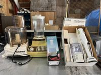 Lot containing four kitchen appliances on industrial table: vintage Cuisinart food processor, vintage blender with glass jar, boxed Braun Handblender with attachments, and a vintage blender jar base