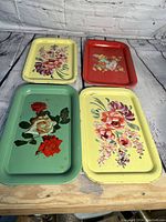 Four vintage metal trays with floral designs in yellow, red, and green backgrounds, showing minor wear and rust spots.