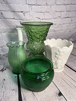 Front view of four vintage glass vases on a wooden surface with a white brick background showing varying shades of green vases and a white milk glass vase with textured patterns.