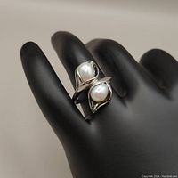 Silver ring on black hand model showing calla lily shaped settings with two white oval pearls.