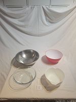 Four mixing bowls arranged separately on a white backdrop: a large stainless steel bowl, a clear glass bowl, a red plastic bowl, and a white plastic bowl with a pour spout.
