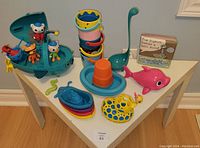 Top view on white small table displaying colorful bath toys including Octonauts submarine with figurines, sensory stacking cups, spotted yellow duck, pink dolphin, various small boats, bath ladle and bath book.