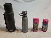 Front view showing all five beverage containers lined up: large black Contigo thermos, gray Contigo water bottle, two colorful kids' Thermos bottles, and their lids.