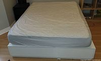 Top-down photo of white Ikea Nordli queen bed frame with visible memory foam mattress and mattress protector. Bed frame shows storage drawers integrated into base.