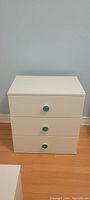Front view of small white dresser showing three drawers each with a green knob.