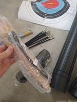 Photo showing 10 black arrows with metal tips and blue plastic fletching, bow string wrapped and wrist/finger guards, multiple archery target sheets, and two black cylindrical plastic quivers.