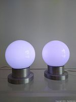 Pair of globe lamps turned on, showing white light glowing from spherical glass diffusers on aluminum bases.