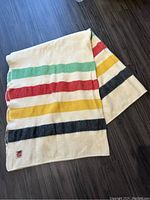 Full view of folded wool blanket showing colorful horizontal stripes in green, red, yellow, and black on cream background.