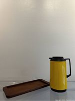 Teak serving tray and yellow insulated pitcher side by side on reflective surface.