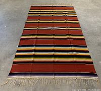 Full view of a large rectangular southwestern-style rug with horizontal colorful stripes and fringed ends laid out on a cement floor.