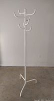 Full view of the white enameled steel coat rack standing upright against a plain wall, showing its structure and three-legged base.