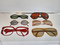 Front view of six vintage sunglasses with various frame colors and lens shades arranged in two rows on a display shelf.