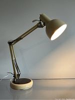 Side view of vintage cream metal adjustable desk lamp lit, showing arm and dome-shaped shade, round base with woodgrain top.