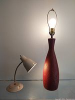 Pair of vintage lamps on a white surface and against a white wall. One lamp is a mid-century modern red ceramic table lamp with a tall base and exposed bulb. The other is a beige goose neck desk lamp with a conical shade and round base.