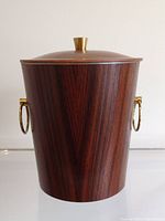 Front view of the cylindrical rosewood ice bucket with brass ring handles and brass knob lid on neutral background.