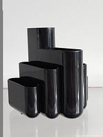 Front angle of the black glossy plastic desk organizer showing its multi-compartment design with tall cylindrical and shorter rectangular sections.