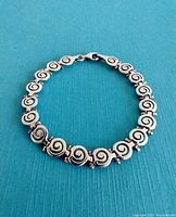 Full view of the bracelet laid flat against a blue textured background, showing the continuous swirl design.