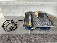 Mastercraft wet tile saw with power cord laid out next to it, showing the full unit on the carpeted floor against a white brick wall.