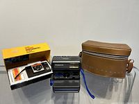 Shows Kodak Instamatic 255 camera with original box, Polaroid camera, and brown leather camera case arranged side by side.